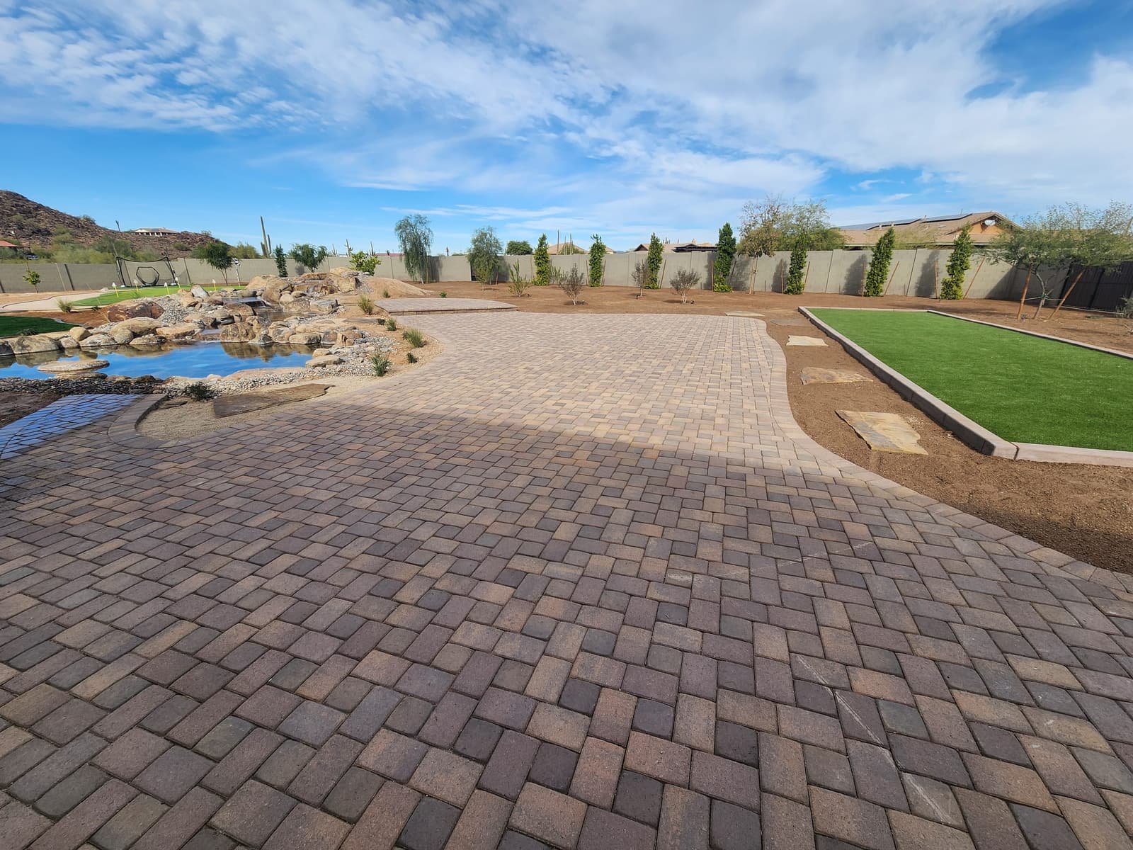 A sweeping paver patio curving past a boulder pond and waterfall toward a turf court and a new row of trees