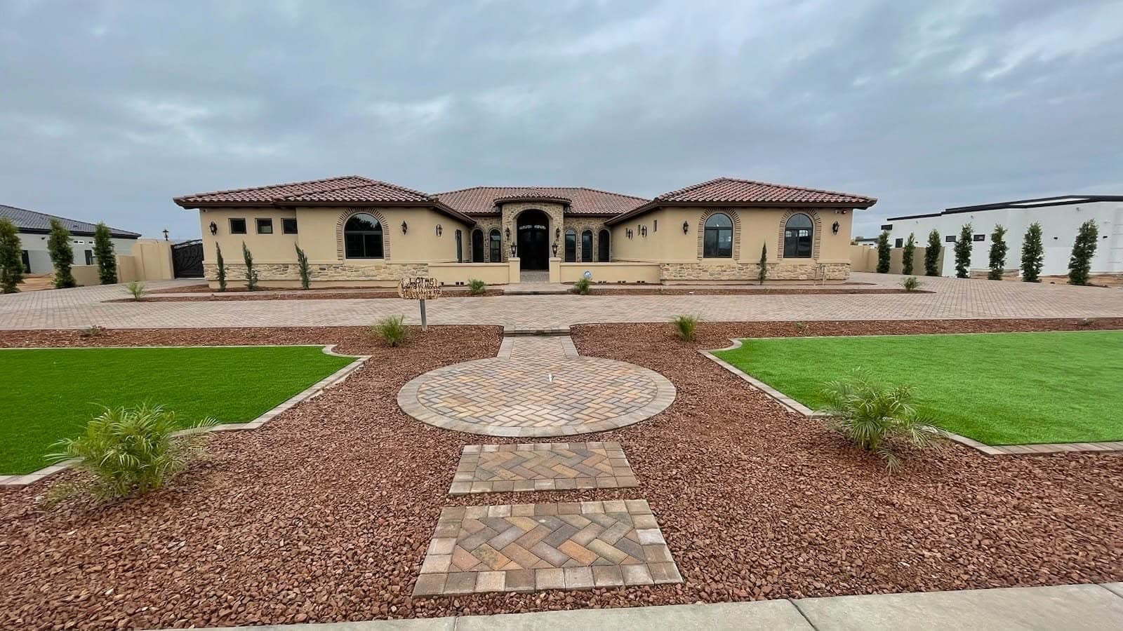 A herringbone paver circle with red rock beds and turf panels in front of a large home