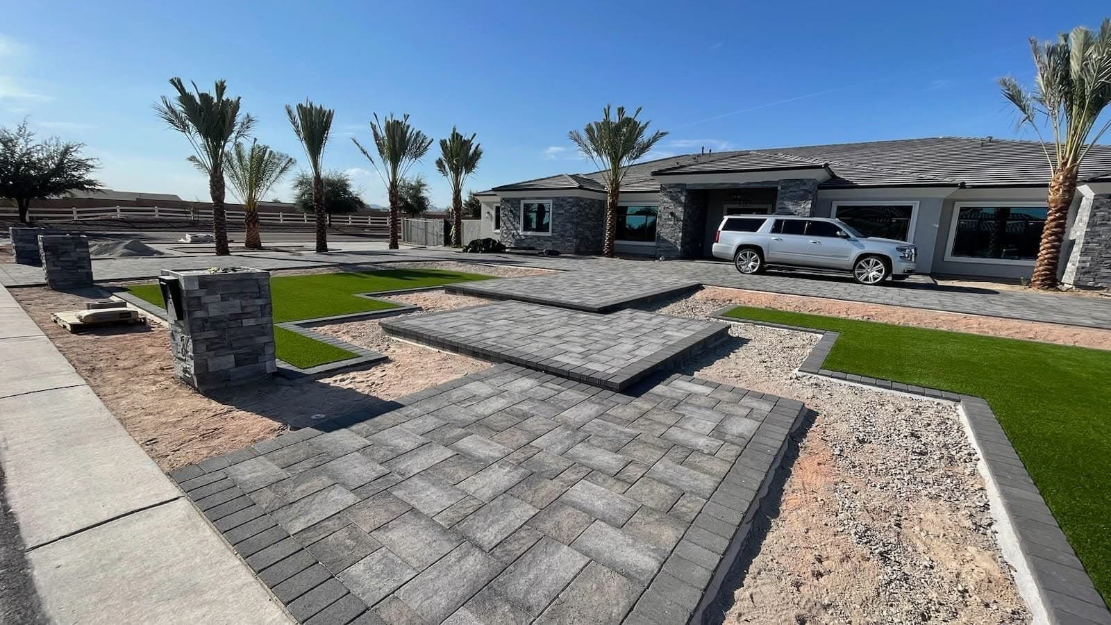 A grey paver drive with stacked-stone columns, turf inlays and mature palms
